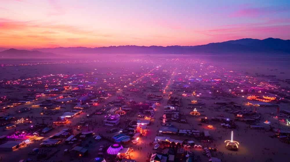 Aerial view of the Burning Man festival in Nevada at sunset, with colorful lights, art installations, and camps spread across the desert under a pink and purple sky.
