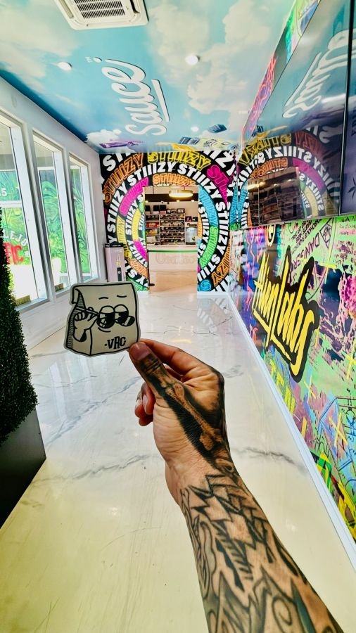 Tattooed hand holding a Vallejo Relief Center sticker inside the dispensary, with colorful graffiti-style wall art, neon brand logos, and a bright hallway leading to the shop counter.