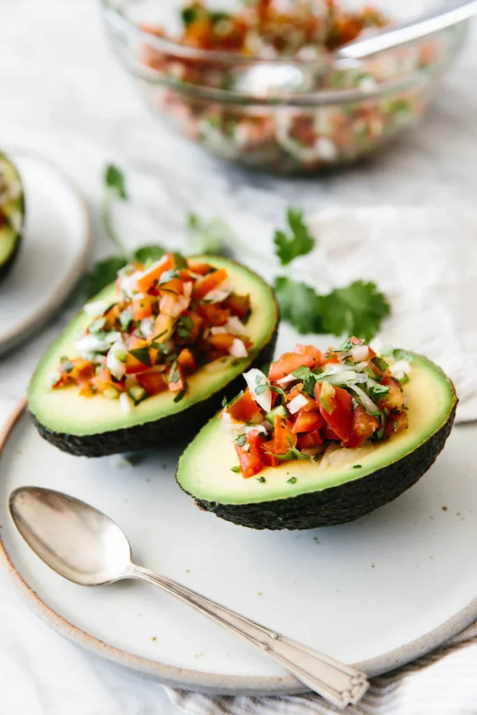 An avocado boat stuffed with Pico de Gallo nicely presented on a white plate on top of a table.
