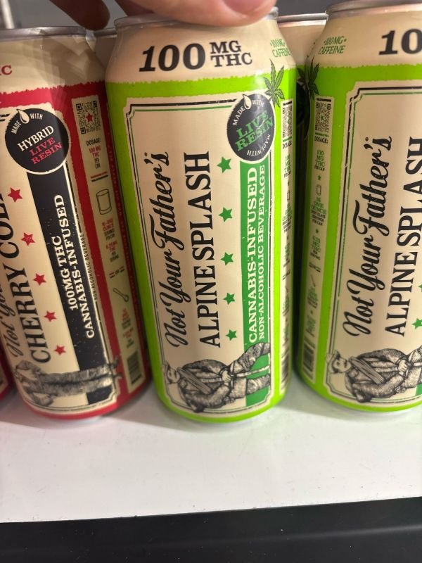 Close-up of “Not Your Father’s” cannabis-infused beverages on a store shelf, featuring Alpine Splash (lemon-lime flavor) and Cherry Cola, each labeled with 100mg THC and infused with live resin.