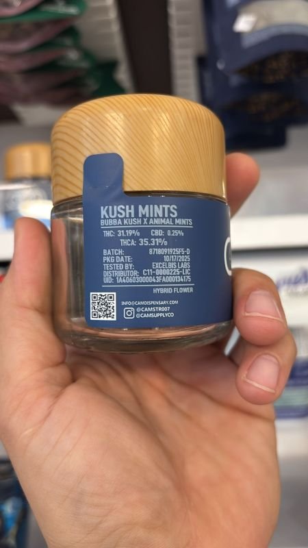 Hand holding a CAM-branded glass jar of Kush Mints hybrid flower, showing detailed strain info including THC content (35.47%), lineage (Bubba Kush x Animal Mints), batch number, and lab testing data.