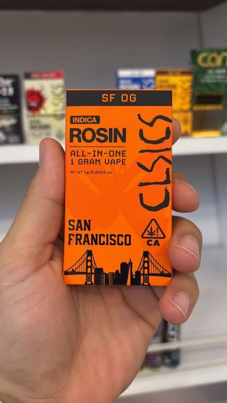 Hand holding a bright orange CLSICS SF OG rosin vape cartridge box labeled 'Indica', 'All-In-One 1 Gram Vape', and 'San Francisco', with cityscape and Golden Gate Bridge artwork.