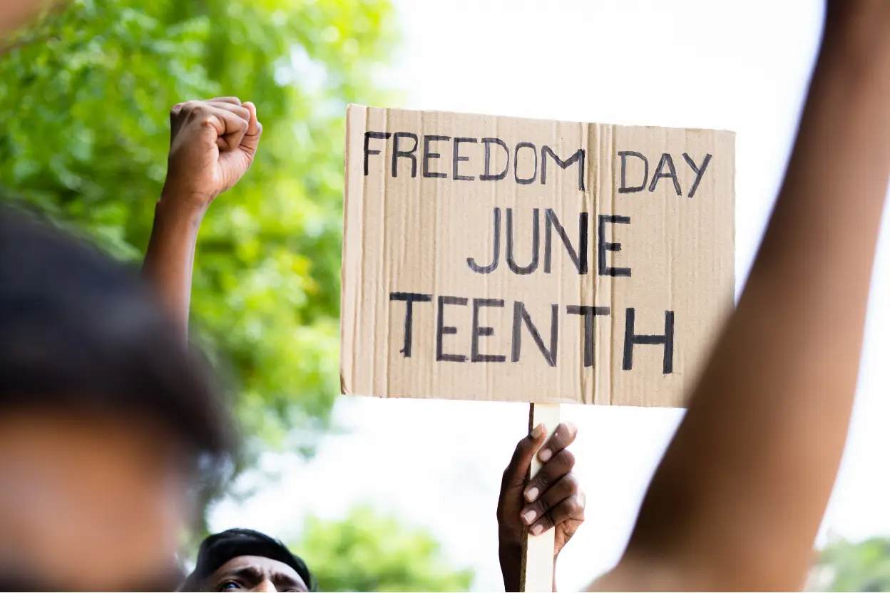 Person holding a cardboard protest sign reading ‘Freedom Day June Teenth’ during a Juneteenth celebration, with raised fists and greenery in the background.