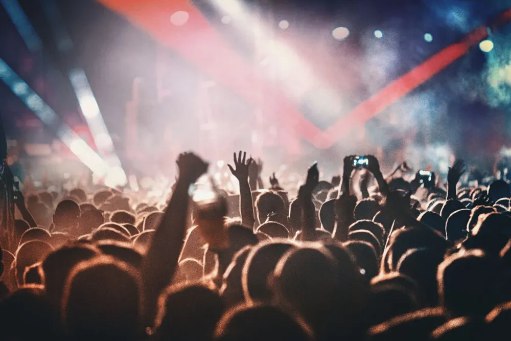 Large crowd at a live concert with hands raised as colorful stage lights shine over the audience.