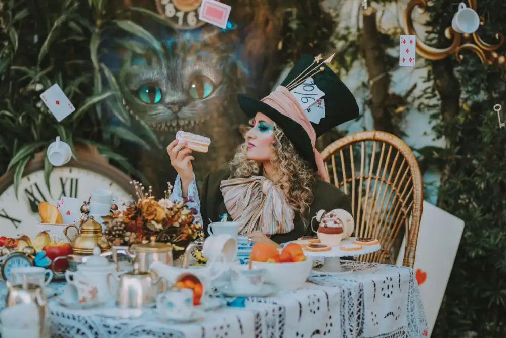 Person dressed as the Mad Hatter seated at an outdoor tea table with desserts, clocks, playing cards, and whimsical decorations.