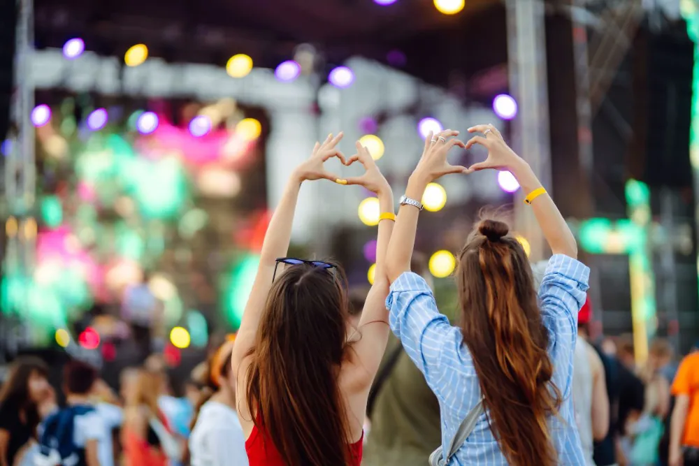 Two people standing in a crowd at a live concert, forming heart shapes with their hands toward a brightly lit stage.