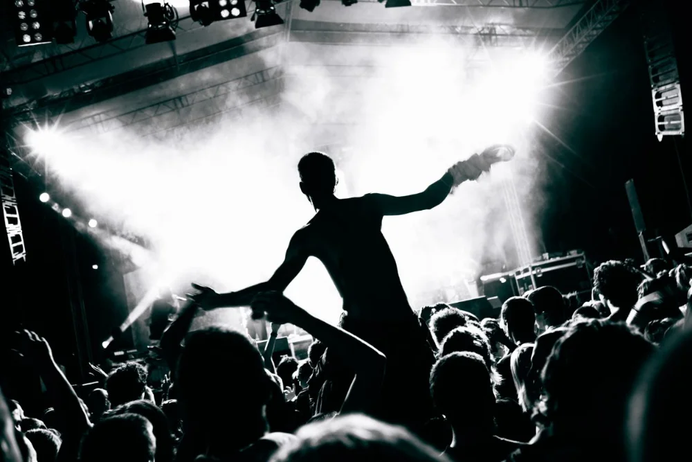 Silhouette of a performer standing above a crowd at a concert, reaching out toward raised hands under bright stage lights.
