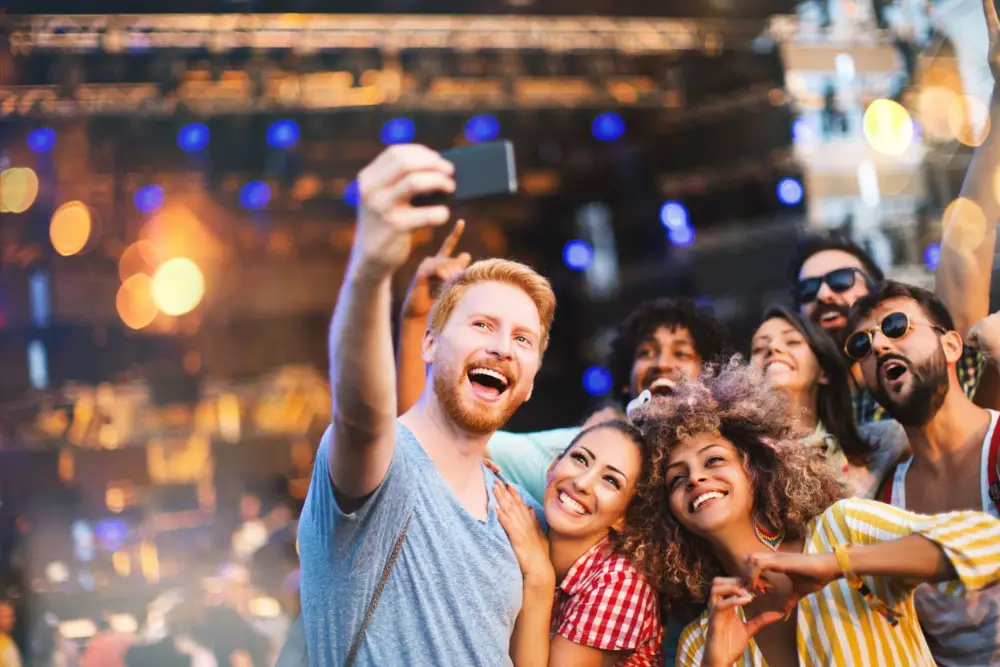 Group of friends smiling and taking a selfie at an outdoor music festival with a stage and lights in the background.