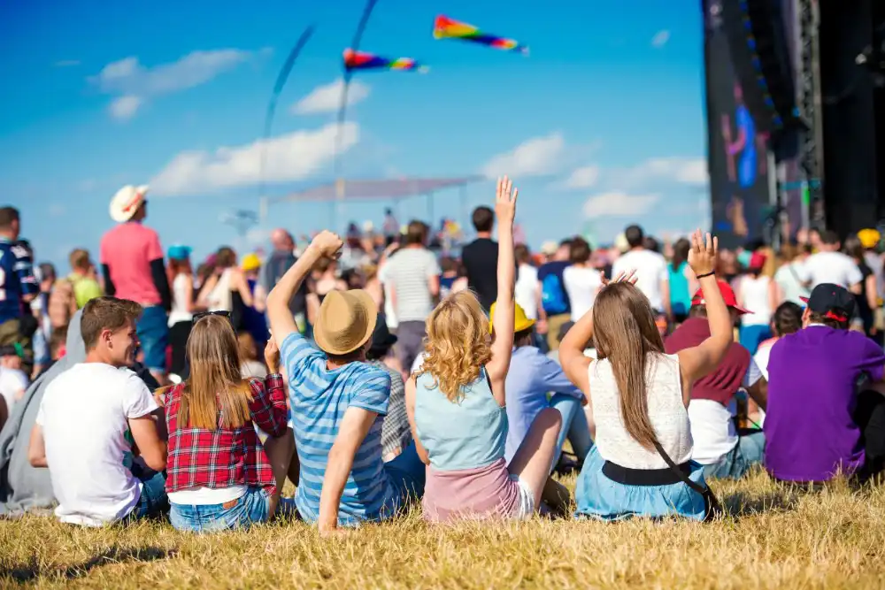 Group of people sitting on grass at an outdoor music festival, raising their arms toward a large stage under a bright blue sky.