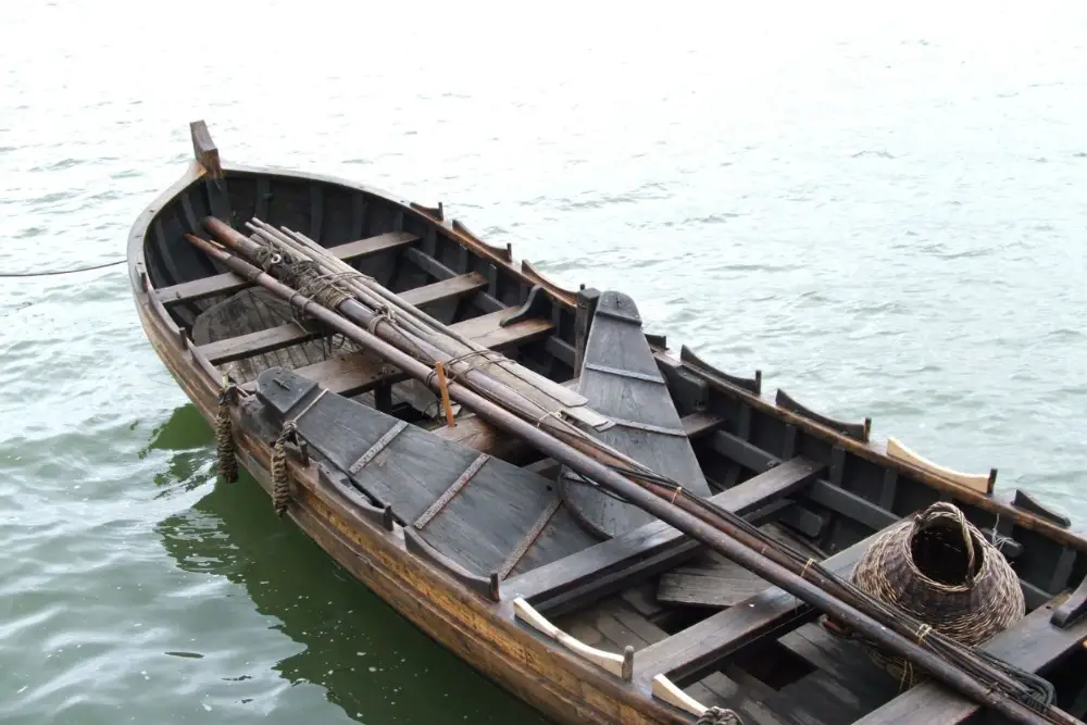 Small wooden whaleboat floating on calm water with oars and coiled rope inside.