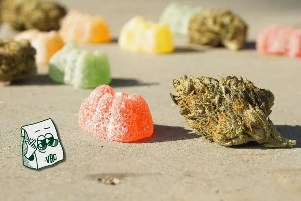 Cannabis gummies and a flower nug on a sandy surface with the VRC mascot bag, representing edible and flower product options at Vallejo Relief Center.