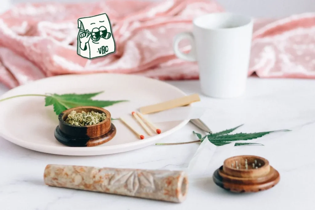 Cannabis flower, pre-roll, and grinder styled in a cozy flat lay with cannabis leaves and VRC mascot bag, evoking a relaxing sesh.